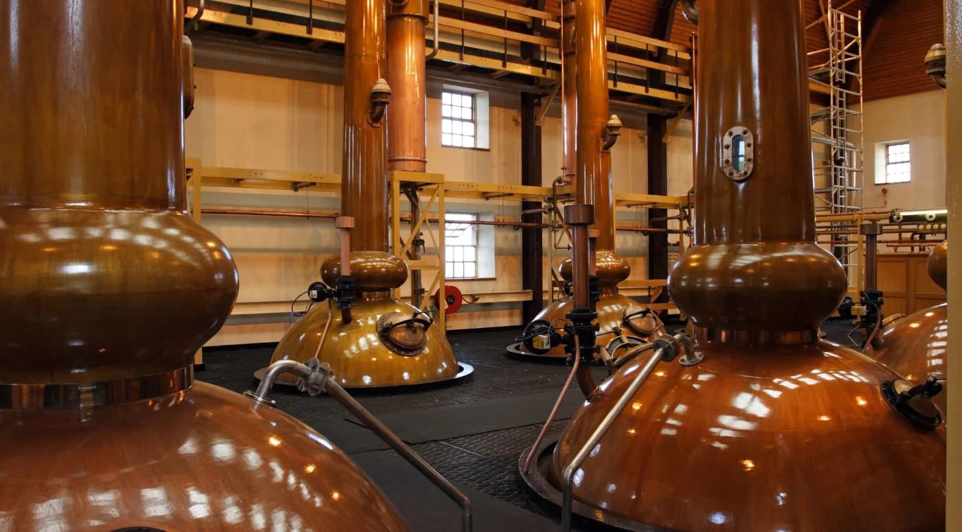 A row of tall copper pot stills in a bright Irish distillery still house